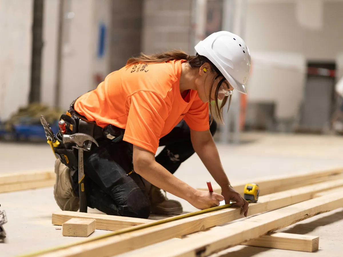 Carpenter | Fast Labour Hire Female construction worker in an orange shirt and hard hat measures a wooden plank with a tape measure on a construction site.