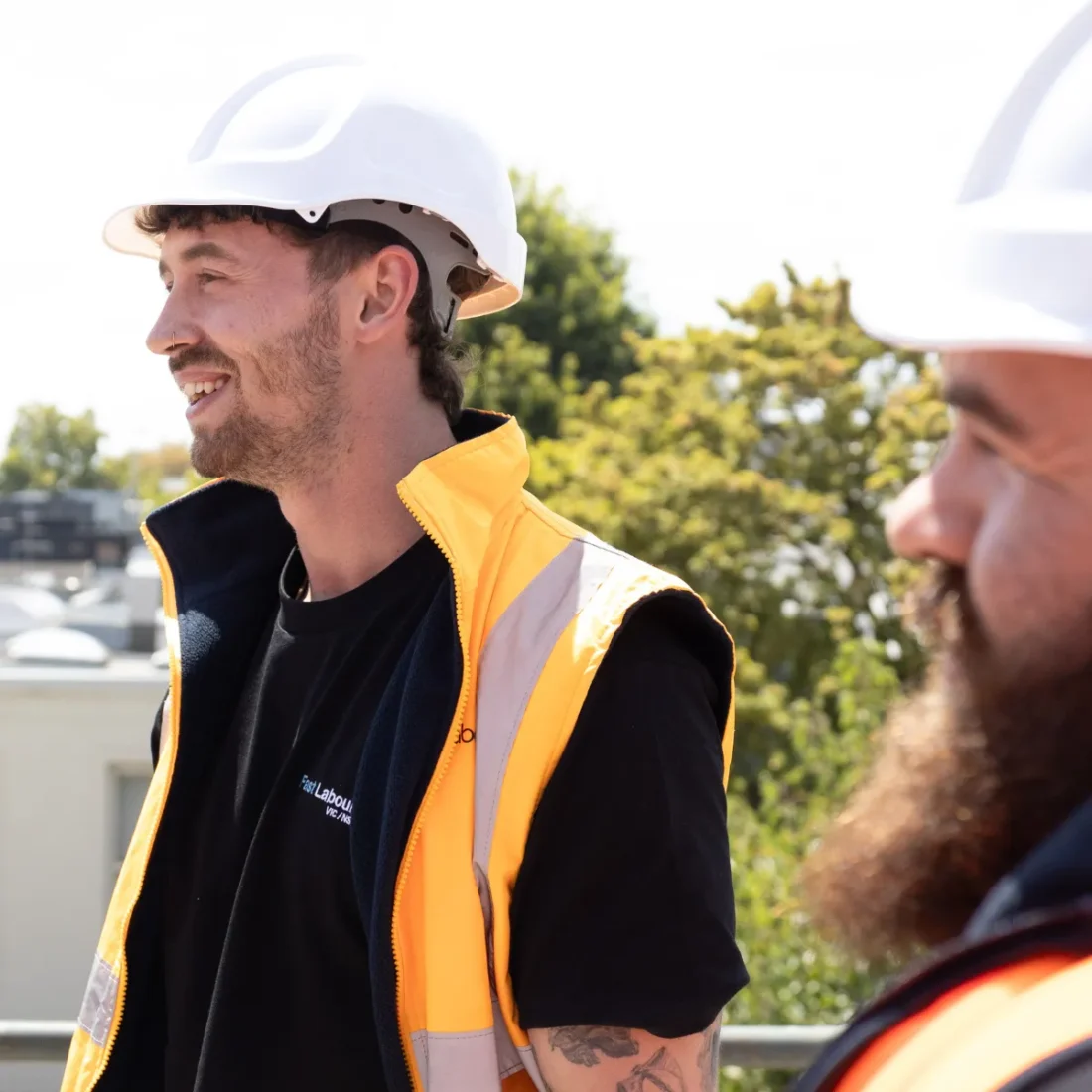 Two construction workers wearing white hard hats and bright orange safety vests talk outdoors on a sunny site.