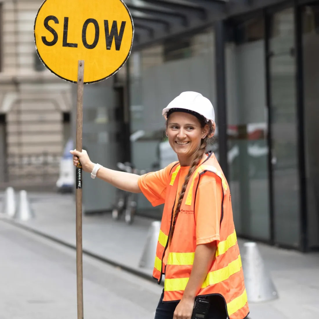 Construction worker in an orange safety vest and white helmet holds a yellow 'SLOW' sign on a city street.