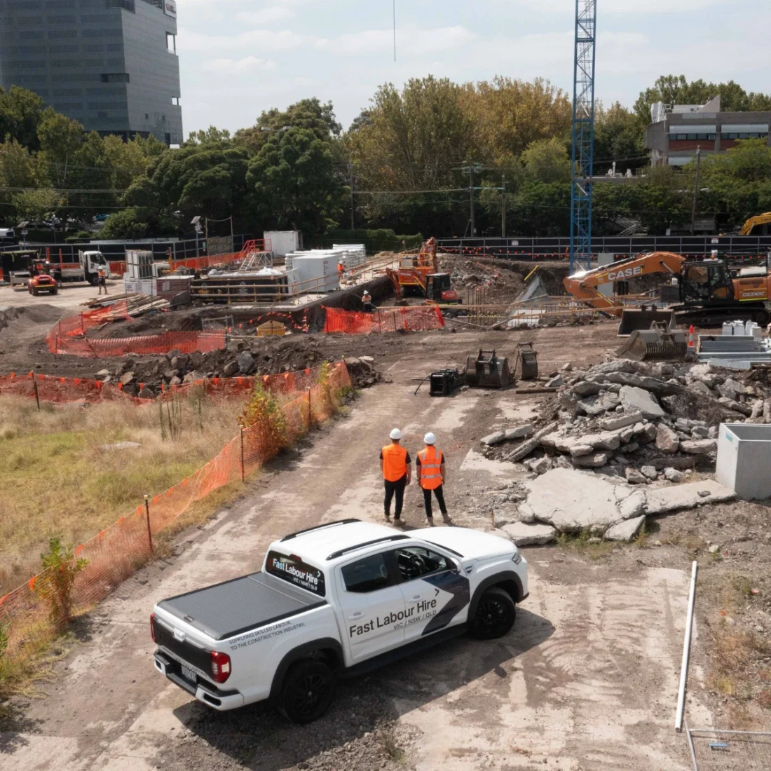 Construction site with two workers in orange vests near a white pickup labeled 'Fast Labour Hire' and orange safety fencing.