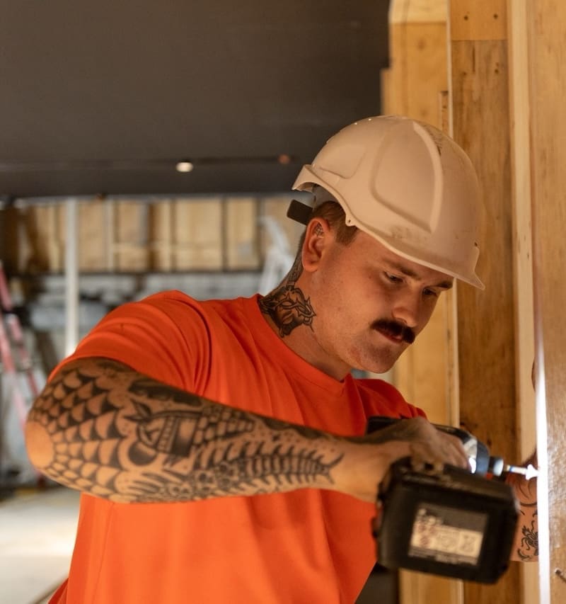 Construction worker in a white hard hat drilling into a wooden stud at a construction site, wearing an orange shirt and visible tattoos on his arm and neck.